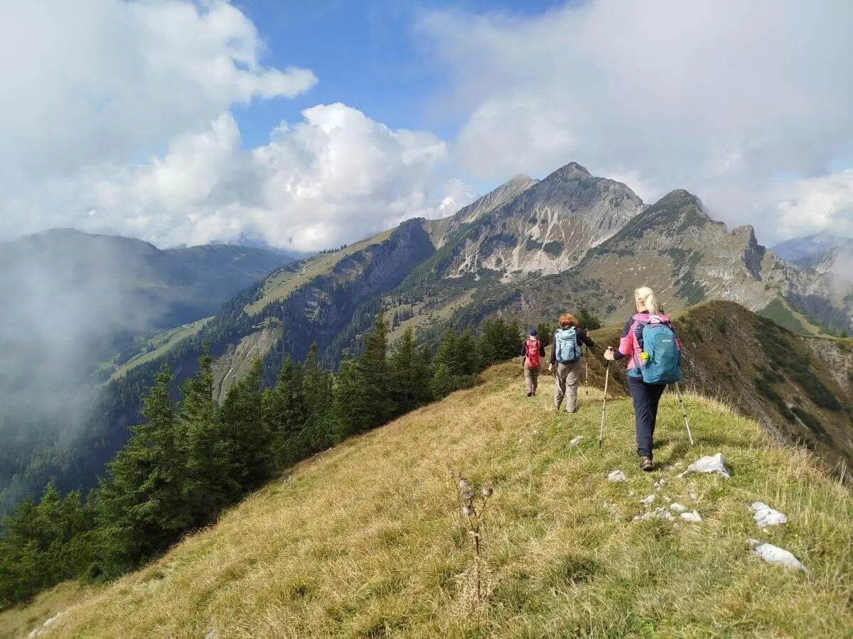 Drei Wandernde stehen vor rustikaler Almhütte aus Holz und Stein; hügelige Landschaft und Wolken im Hintergrund. | © DAV Markt Schwaben | Foto Helga Peters