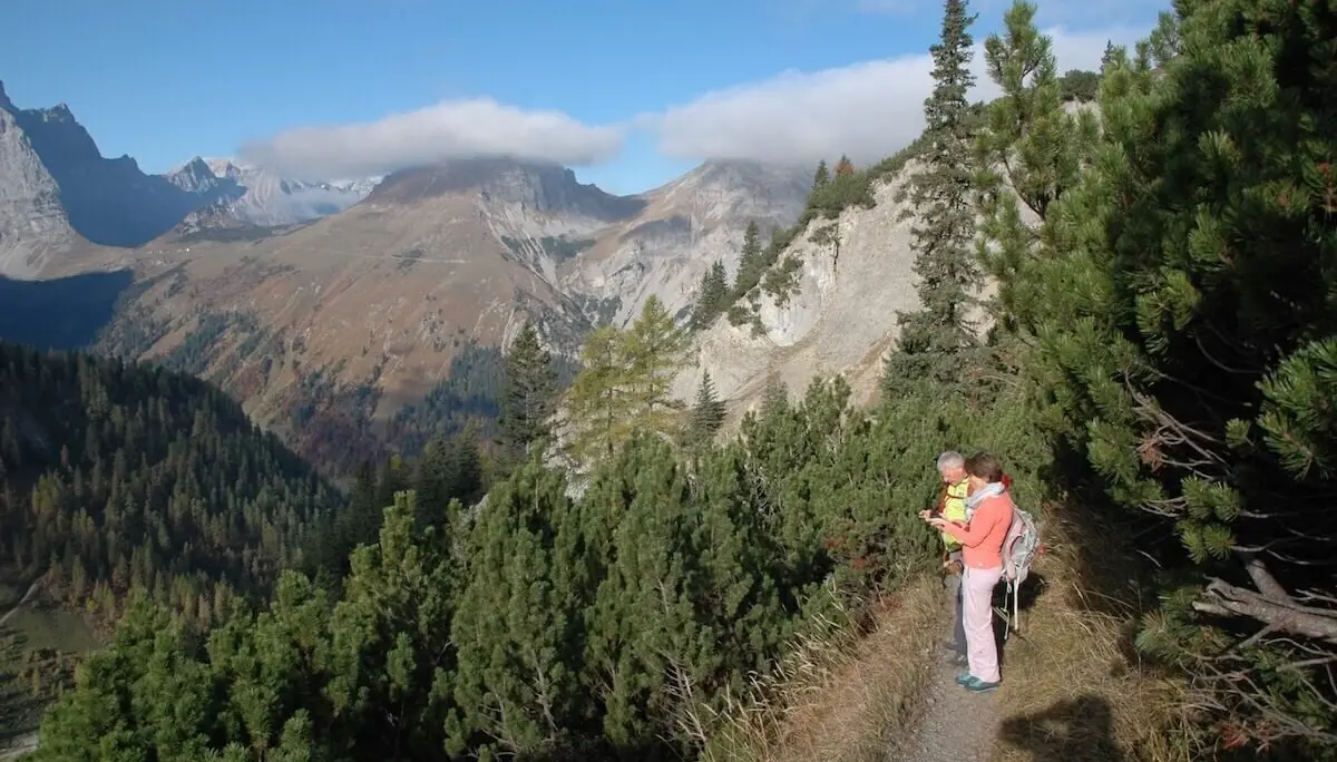 Zwei Wandernde auf einem schmalen Pfad durch dichte Nadelbäume; im Hintergrund steile Bergflanken und Wolken. | © DAV Markt Schwaben · Foto: Matzinger