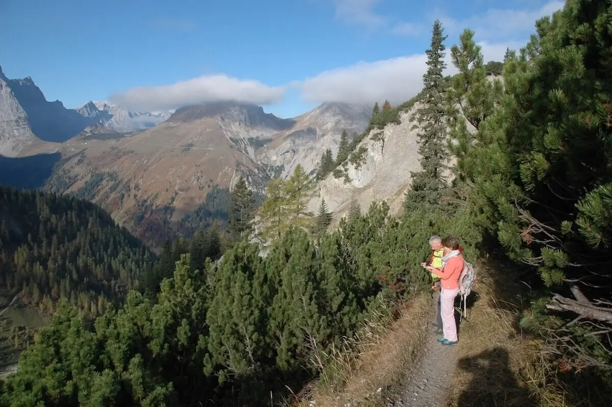 Zwei Wandernde auf einem schmalen Pfad durch dichte Nadelbäume; im Hintergrund steile Bergflanken und Wolken. | © DAV Markt Schwaben · Foto: Matzinger