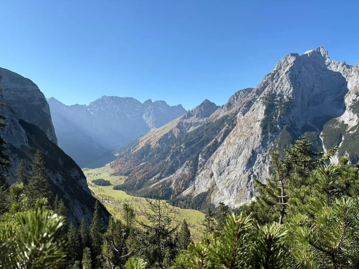 Grüne Nadelbäume im Vordergrund rahmen ein tiefes Tal mit Wiesen und Felsen; dahinter steile Bergflanken bei Sonnenschein. | © DAV Markt Schwaben · Foto: Markus Zimmermann