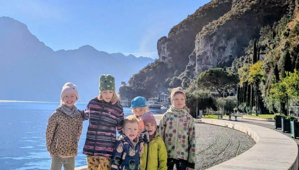 Mehrere Erwachsene und Kinder sitzen auf Matten und Baumstämmen in einem herbstlichen Waldstück. Ein Kind steht in der Mitte mit erhobenen Armen. Alle tragen warme Kleidung, im Hintergrund sind bunte Blätter, ein Zaun und Berge zu sehen. | © DAV Markt Schwaben · Foto: Silke Titze