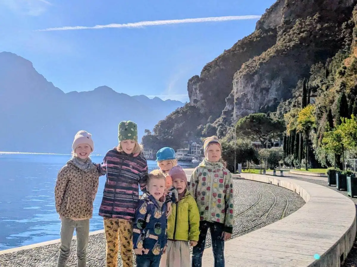 Mehrere Erwachsene und Kinder sitzen auf Matten und Baumstämmen in einem herbstlichen Waldstück. Ein Kind steht in der Mitte mit erhobenen Armen. Alle tragen warme Kleidung, im Hintergrund sind bunte Blätter, ein Zaun und Berge zu sehen. | © DAV Markt Schwaben · Foto: Silke Titze