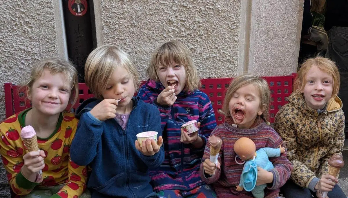 Drei Kinder in Kletterausrüstung stehen auf einer Wiese neben einer schwarzen Ziege. Ein Kind hält einen Ast, vermutlich zum Füttern. Im Hintergrund klettern weitere Personen an einem Felsen, umgeben von Bäumen und Sonnenlicht. | © DAV Markt Schwaben · Foto: Silke Titze