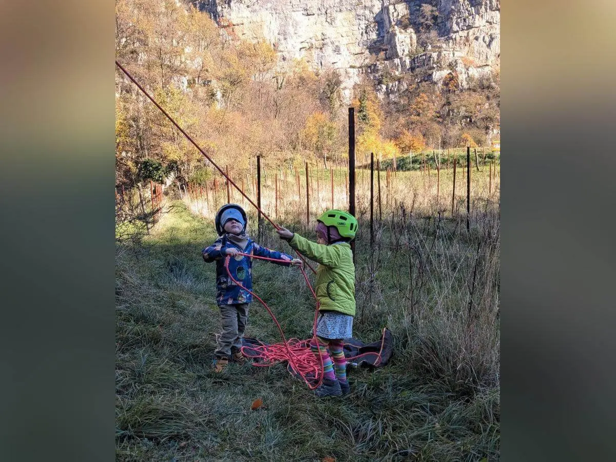 Ein Kind mit rotem Helm und Kletterausrüstung steigt eine steile Felswand hinauf. Dahinter klettert eine weitere Person mit blauer Ausrüstung. Beide sind gesichert und konzentriert in Bewegung. Die Szene spielt sich an einem sonnigen Felsen in der Natur ab. | © DAV Markt Schwaben · Foto: Silke Titze