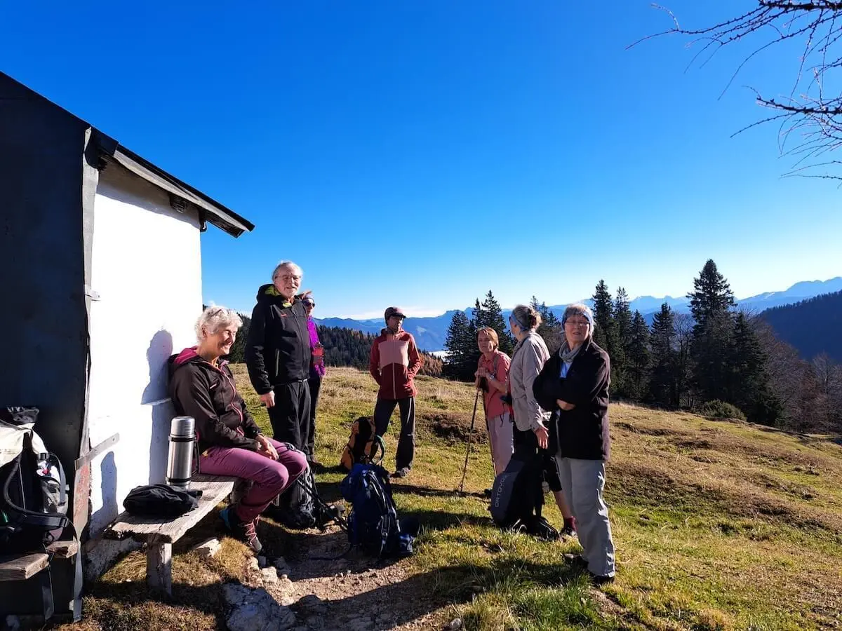 Sieben Wandernde machen Rast vor einer kleinen weißen Hütte mit Bank. Einige stehen, eine Person sitzt, Rucksäcke und Thermosflasche liegen bereit. Im Hintergrund sind bewaldete Hügel und ferne Berggipfel unter klarem Himmel zu sehen – die Szene wirkt ruhig und gemeinschaftlich. | © DAV Markt Schwaben | Foto: Tanja Döhler