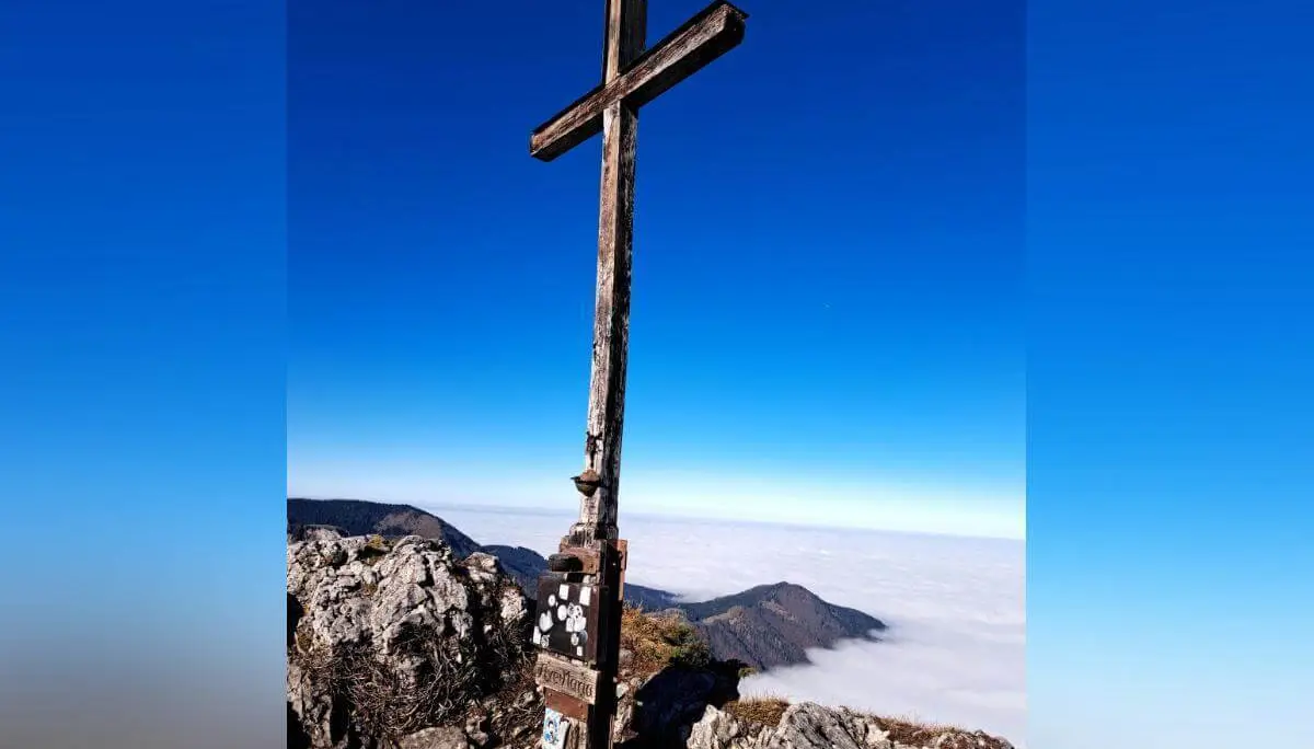 Ein großes Holzkreuz steht auf einem felsigen Gipfel mit Blick auf Bergketten und Wolkenmeer. Am Sockel sind Plaketten und die Jahreszahl 1927 zu sehen, die Höhe ist mit 2141 Metern angegeben. Die Szene vermittelt Weite, Symbolkraft und alpine Erhabenheit bei klarem Wetter. | © DAV Markt Schwaben | Foto: Tanja Döhler
