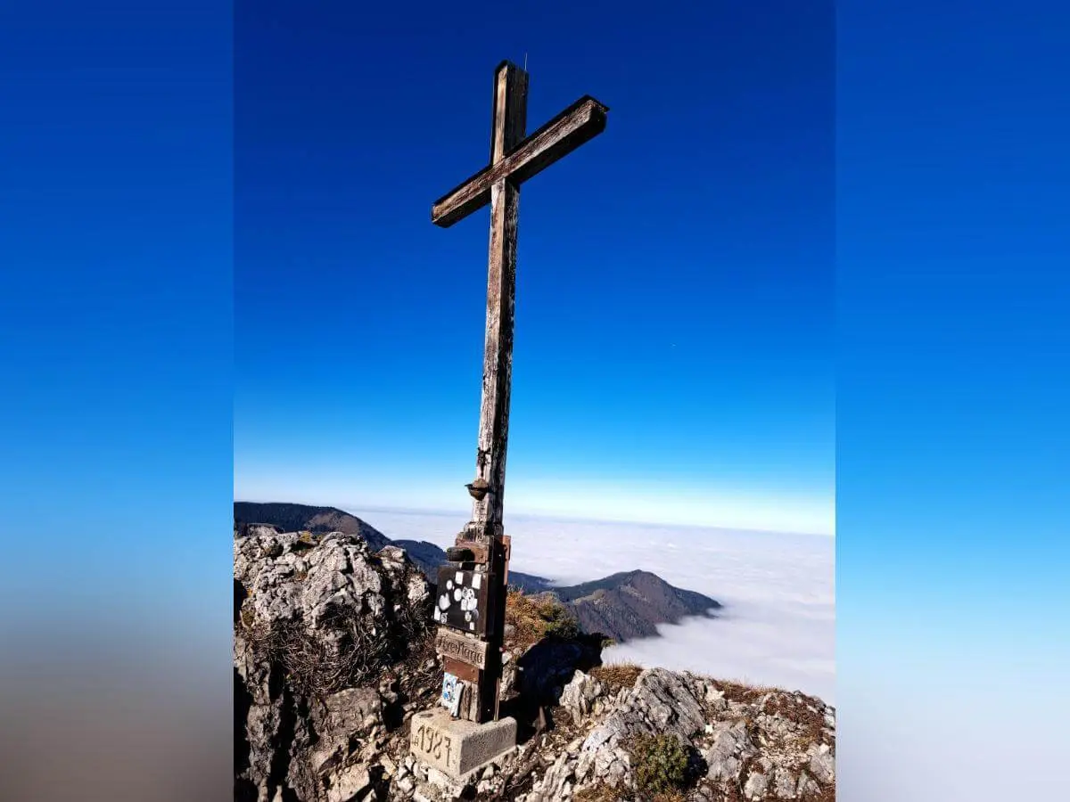 Ein großes Holzkreuz steht auf einem felsigen Gipfel mit Blick auf Bergketten und Wolkenmeer. Am Sockel sind Plaketten und die Jahreszahl 1927 zu sehen, die Höhe ist mit 2141 Metern angegeben. Die Szene vermittelt Weite, Symbolkraft und alpine Erhabenheit bei klarem Wetter. | © DAV Markt Schwaben | Foto: Tanja Döhler