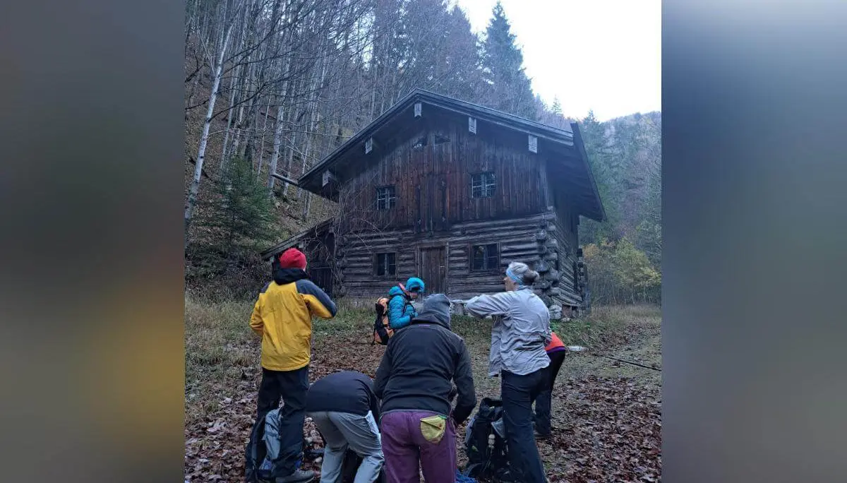 Vor einer rustikalen Holzhütte im Wald versammelt sich eine Wandergruppe, umgeben von herbstlichem Laub. Die Szene wirkt ruhig und stimmungsvoll, leichter Nebel liegt über dem Boden und verleiht der Umgebung eine geheimnisvolle Atmosphäre. Die Kleidung der Personen deutet auf eine Pause während einer herbstlichen Tour hin. | © DAV Markt Schwaben | Foto: Tanja Döhler