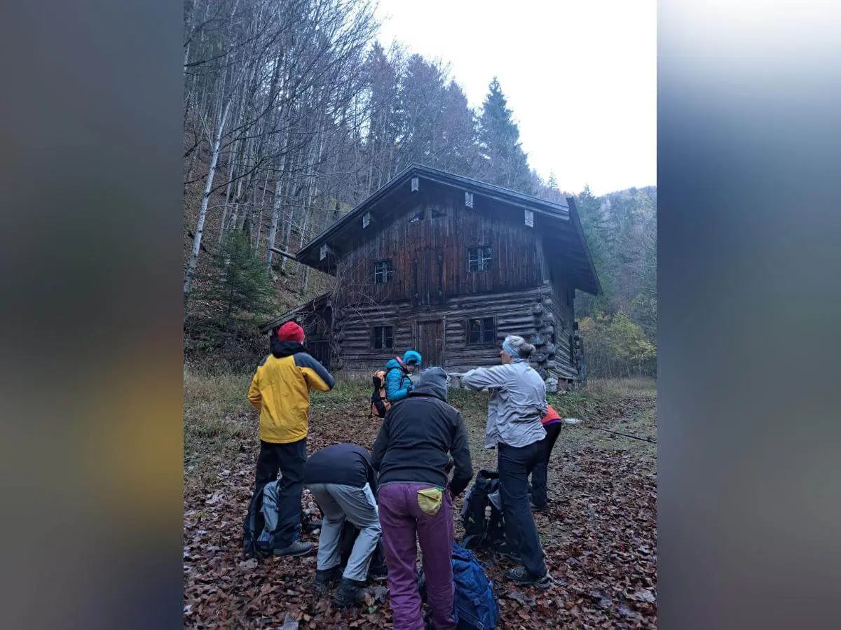 Vor einer rustikalen Holzhütte im Wald versammelt sich eine Wandergruppe, umgeben von herbstlichem Laub. Die Szene wirkt ruhig und stimmungsvoll, leichter Nebel liegt über dem Boden und verleiht der Umgebung eine geheimnisvolle Atmosphäre. Die Kleidung der Personen deutet auf eine Pause während einer herbstlichen Tour hin. | © DAV Markt Schwaben | Foto: Tanja Döhler