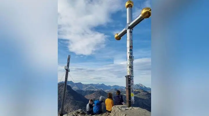 Vier Personen sitzen auf dem Gipfel des Kranzhorns vor einem großen Gipfelkreuz und blicken auf die umliegenden Berge. Das Bild zeigt Gemeinschaft und Naturerlebnis bei einer Familienwanderung. | © DAV Markt Schwaben | Foto: Maria Wenisch