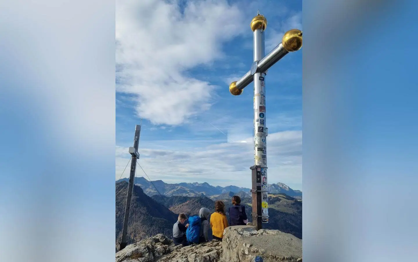 Vier Personen sitzen auf dem Gipfel des Kranzhorns vor einem großen Gipfelkreuz und blicken auf die umliegenden Berge. Das Bild zeigt Gemeinschaft und Naturerlebnis bei einer Familienwanderung. | © DAV Markt Schwaben | Foto: Maria Wenisch