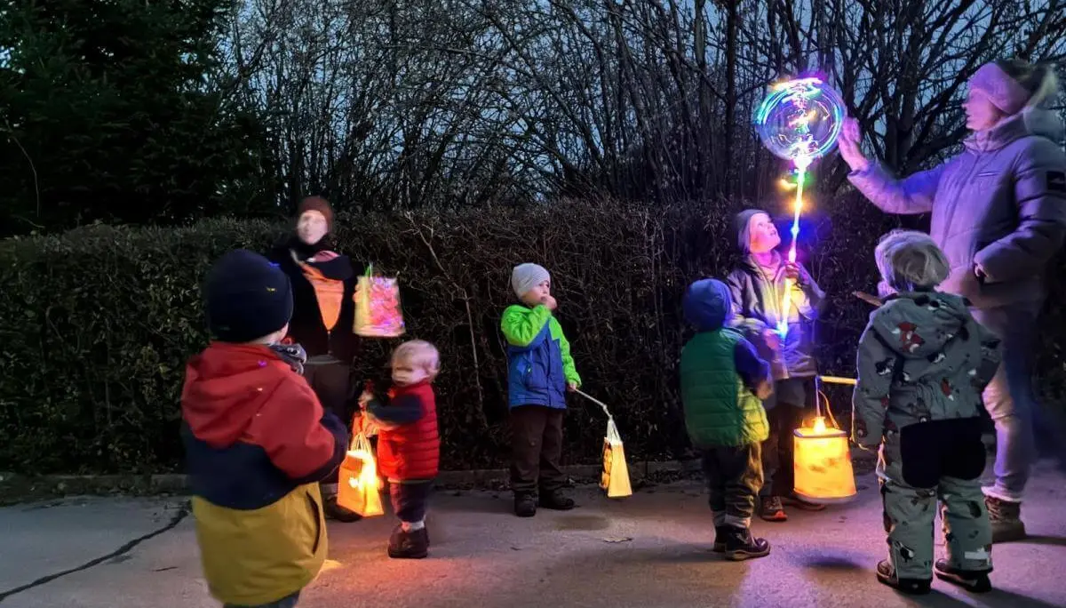 Mehrere Kinder und Erwachsene stehen draußen bei Dämmerung auf einem gepflasterten Platz. Die Kinder halten leuchtende Laternen, ein Erwachsener trägt einen bunten Leuchtstab. Alle sind warm angezogen, im Hintergrund sind Hecken und Bäume zu sehen. | © DAV Markt Schwaben · Foto: Lara Wolff
