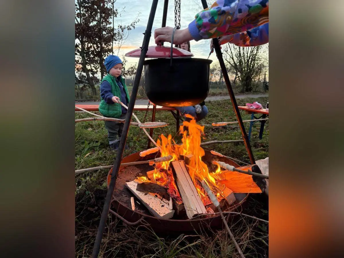 Ein Kind mit warmer Kleidung steht neben einem Lagerfeuer und hält einen Stock ins Feuer. Ein Erwachsener hebt den Deckel eines schwarzen Topfs, der über dem Feuer hängt. Rundherum liegen Holzscheite, im Hintergrund sind Bäume und Sitzbänke sichtbar. | © DAV Markt Schwaben · Foto: Lara Wolff