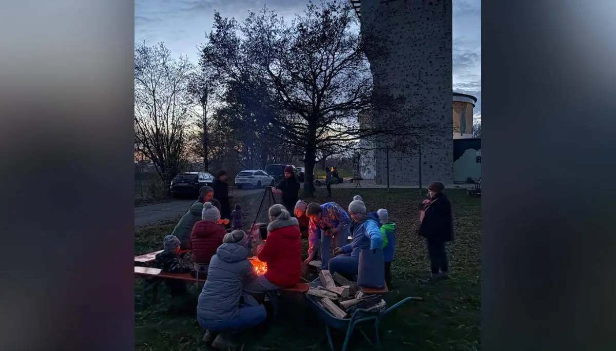 Mehrere Personen sitzen bei Dämmerung um ein Lagerfeuer, warm gekleidet mit Jacken und Mützen. Sie unterhalten sich oder schauen ins Feuer. Im Hintergrund stehen ein Kletterturm, ein rundes Gebäude und geparkte Autos. Die Bäume sind kahl, es wirkt herbstlich. | © DAV Markt Schwaben · Foto: Lara Wolff