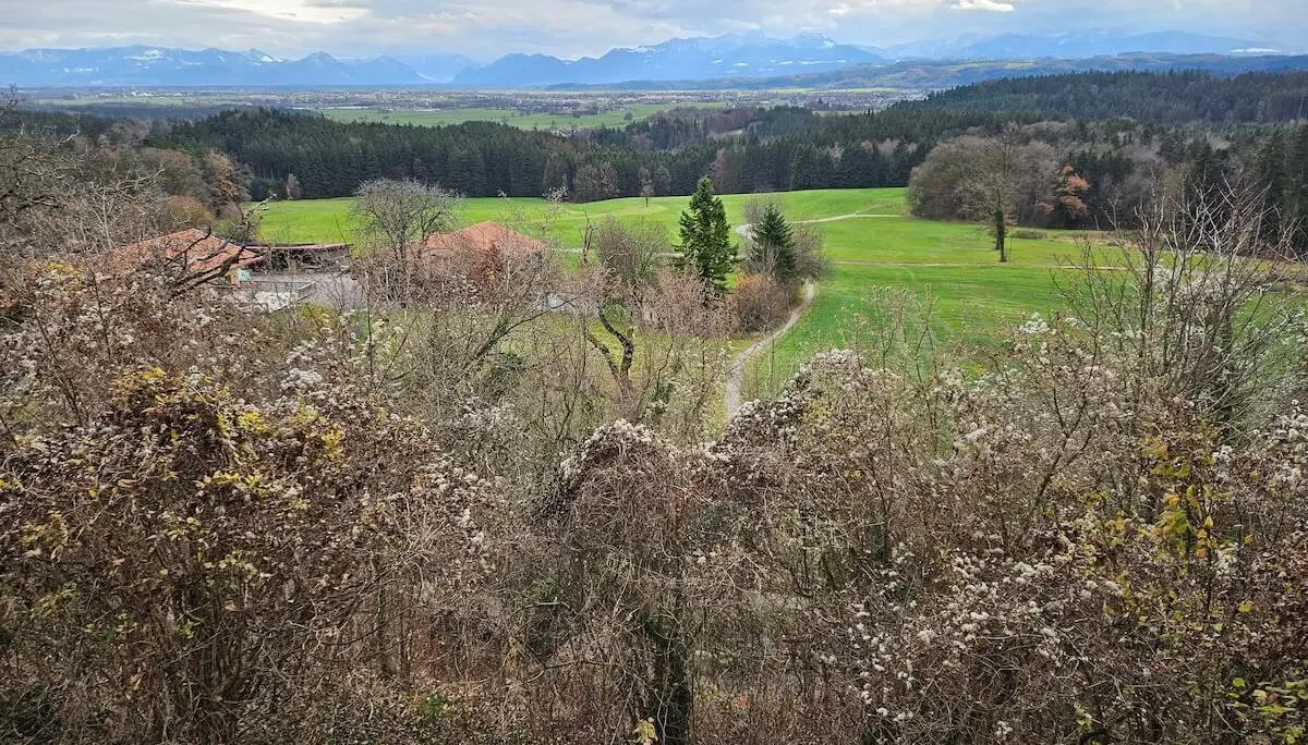 Vorne Sträucher und Bauernhof, mittig Wiesen mit Weg, hinten Wald und schneebedeckte Berge unter Wolken. | © DAV Markt Schwaben · Foto: Hubert Inhofer