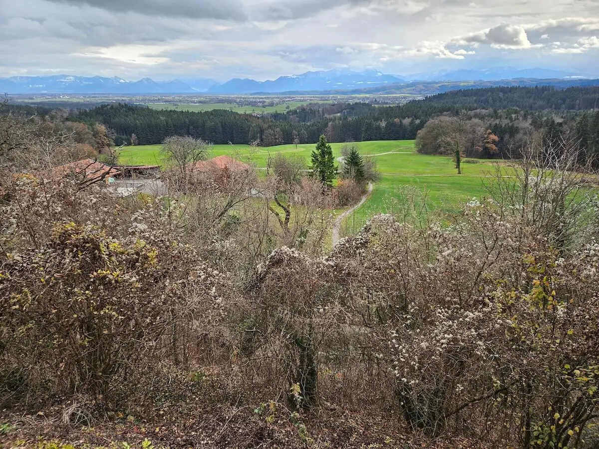 Vorne Sträucher und Bauernhof, mittig Wiesen mit Weg, hinten Wald und schneebedeckte Berge unter Wolken. | © DAV Markt Schwaben · Foto: Hubert Inhofer