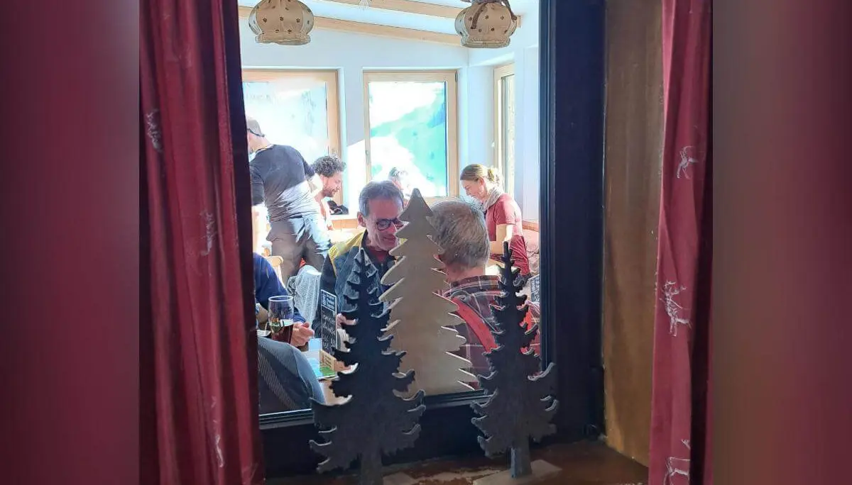 Blick durch Fenster mit roten Vorhängen auf hellen Gastraum, Personen sitzen und unterhalten sich, Deko im Vordergrund. | © DAV Markt Schwaben · Foto Tanja Döhler