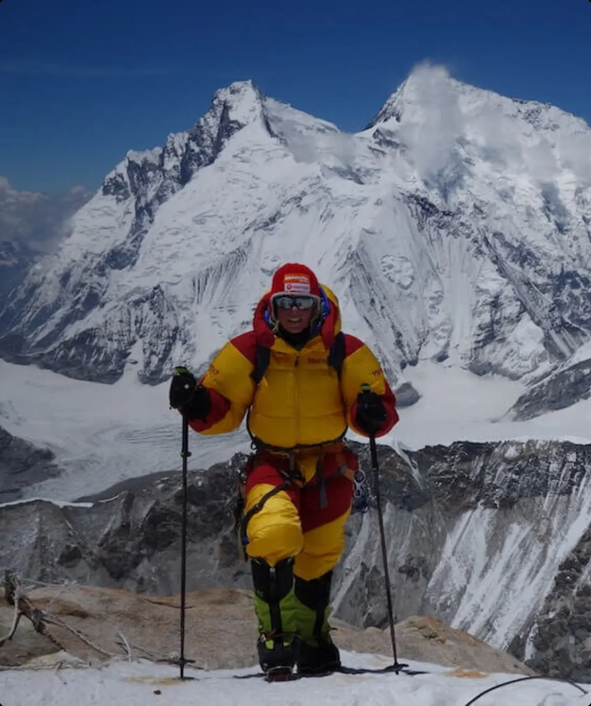 Ein Bergsteiger in gelbem und rotem Anzug steht mit Kletterausrüstung auf einem schneebedeckten Bergrücken, umgeben von Gipfeln. | © Alix von Melle