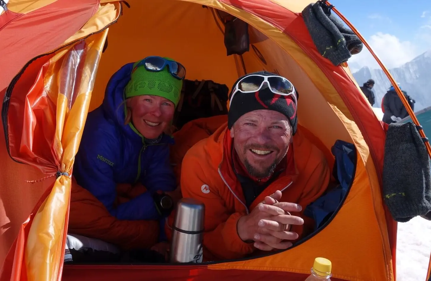 Zwei Personen sitzen in einem orangefarbenen Zelt auf einem verschneiten Berg, tragen Winterausrüstung und Sonnenbrillen. | © Alix von Melle