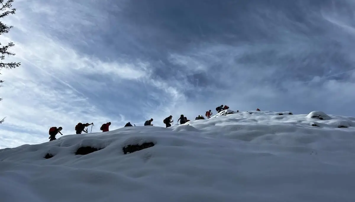 Eine Gruppe von Wandernden steigt mit Rucksäcken und Stöcken einen verschneiten Hang hinauf. Der Himmel ist teils bewölkt, die Personen sind als Silhouetten vor der dramatischen Wolkenkulisse sichtbar. | © DAV Markt Schwaben · Foto Markus Sellmeier
