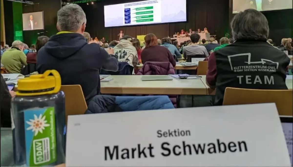 Delegierte der DAV-Sektion Markt Schwaben sitzen im Konferenzsaal der Hauptversammlung in Passau. Im Vordergrund: Namensschild der Sektion und DAV-Wasserflasche. Auf der Bühne läuft eine Präsentation, Teilnehmende verfolgen konzentriert das Geschehen. | © DAV Markt Schwaben · Fotograf unbekannt