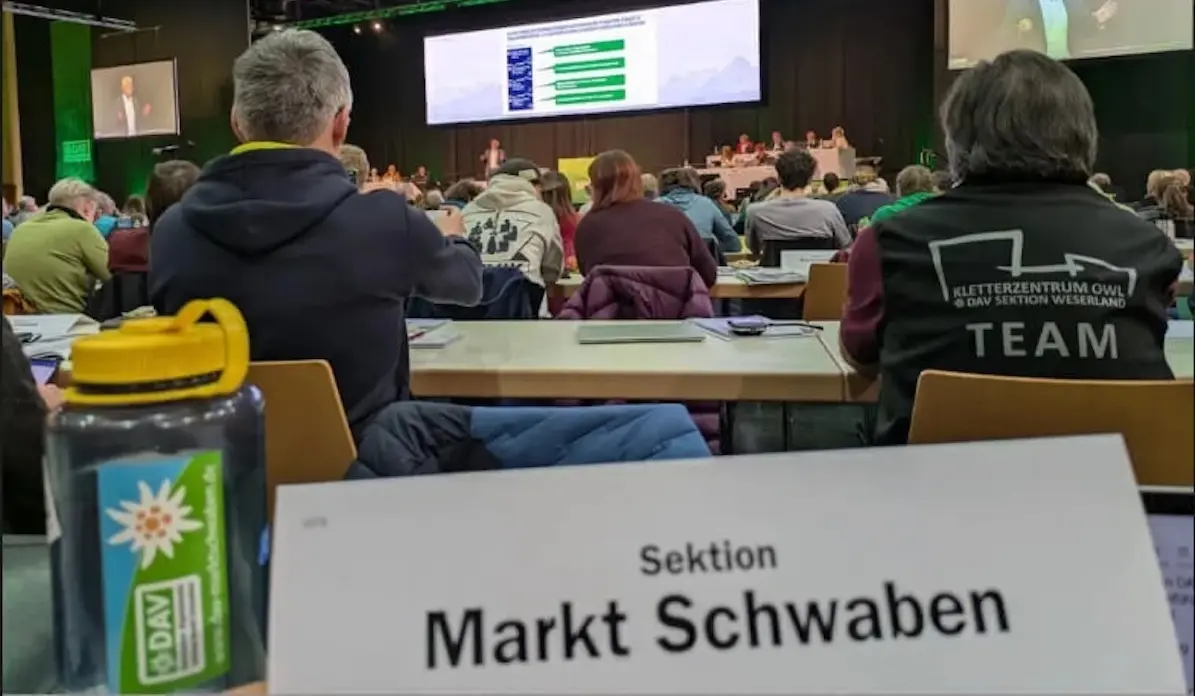 Delegierte der DAV-Sektion Markt Schwaben sitzen im Konferenzsaal der Hauptversammlung in Passau. Im Vordergrund: Namensschild der Sektion und DAV-Wasserflasche. Auf der Bühne läuft eine Präsentation, Teilnehmende verfolgen konzentriert das Geschehen. | © DAV Markt Schwaben · Fotograf unbekannt