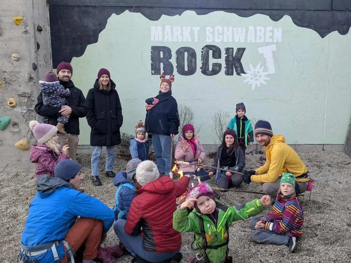 Kinder und Erwachsene stehen vor bemalter Kletterwand mit Feuerstelle; Winterkleidung, lockere Gruppe, Dämmerung. | © DAV Markt Schwaben · Familiengruppe