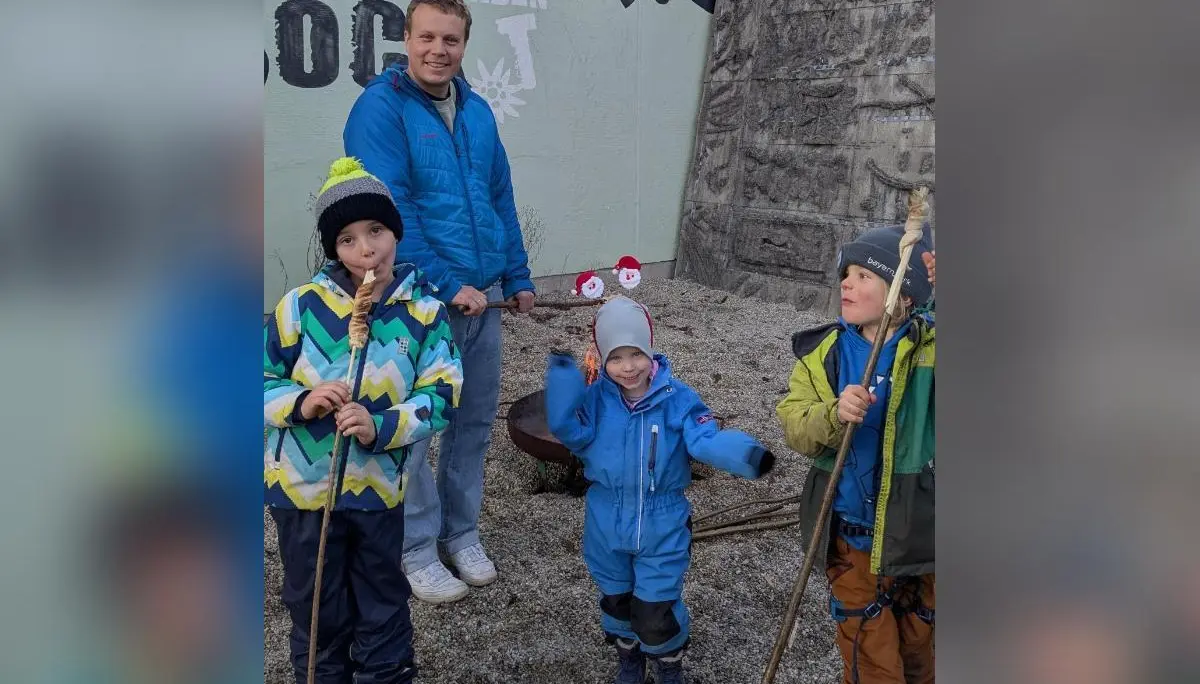 Vier Kinder mit Stöcken vor bemalter Wand und Feuerstelle; Winterkleidung, Erwachsene im Hintergrund, Kiesplatz. | © DAV Markt Schwaben · Familiengruppe