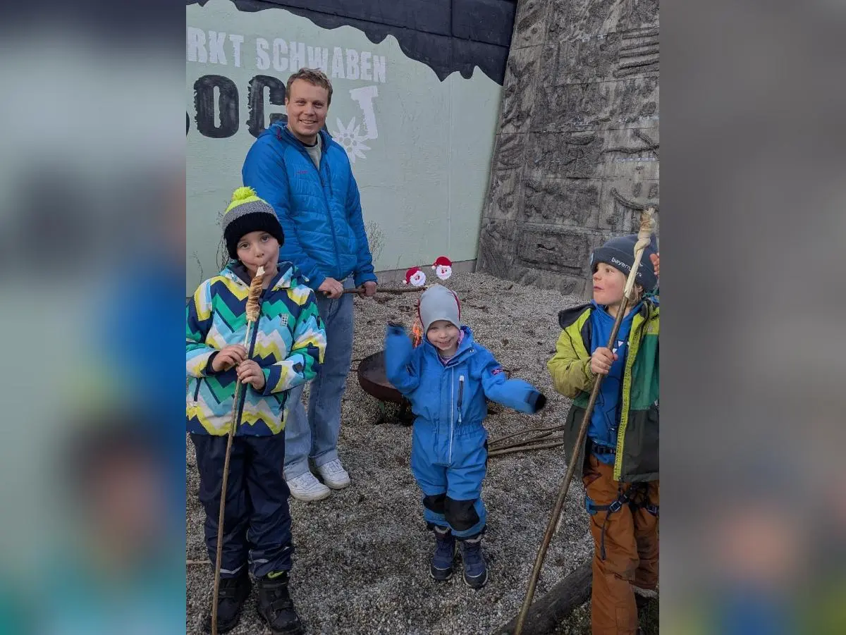 Vier Kinder mit Stöcken vor bemalter Wand und Feuerstelle; Winterkleidung, Erwachsene im Hintergrund, Kiesplatz. | © DAV Markt Schwaben · Familiengruppe