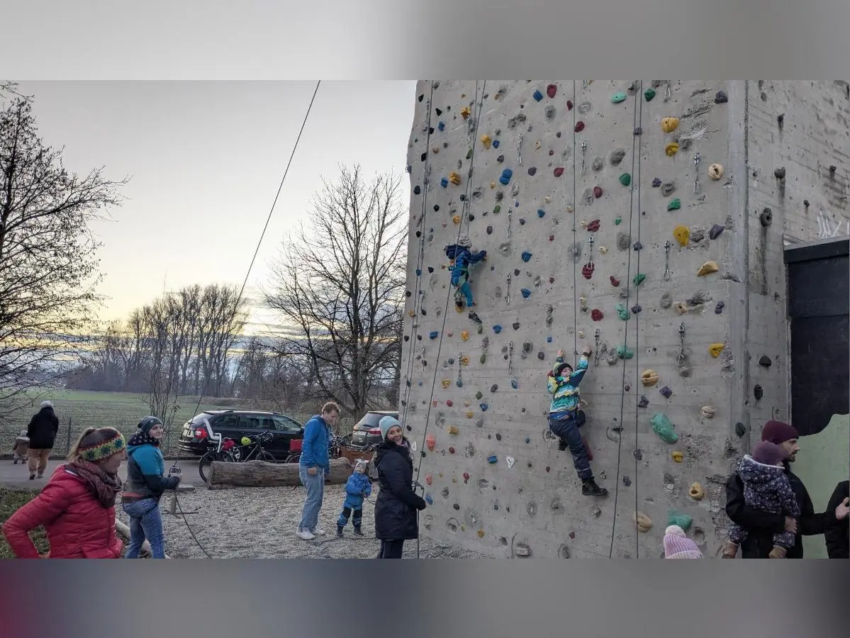 Zwei Kinder klettern gesichert an der bunten Kletterwand; Erwachsene stehen daneben, Bäume im Hintergrund. | © DAV Markt Schwaben · Familiengruppe