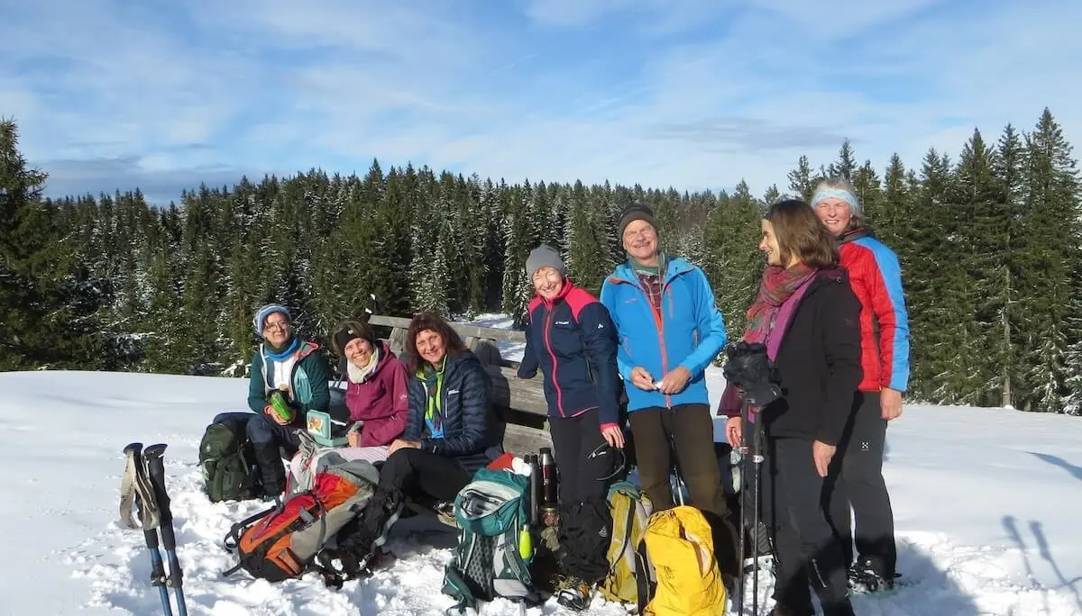 Sieben Wandernde pausieren an einer Holzbank im Schnee; im Hintergrund verschneite Tannen und blauer Himmel mit Wolken. | © DAV Markt Schwaben | Foto: Gerlinde Hübl