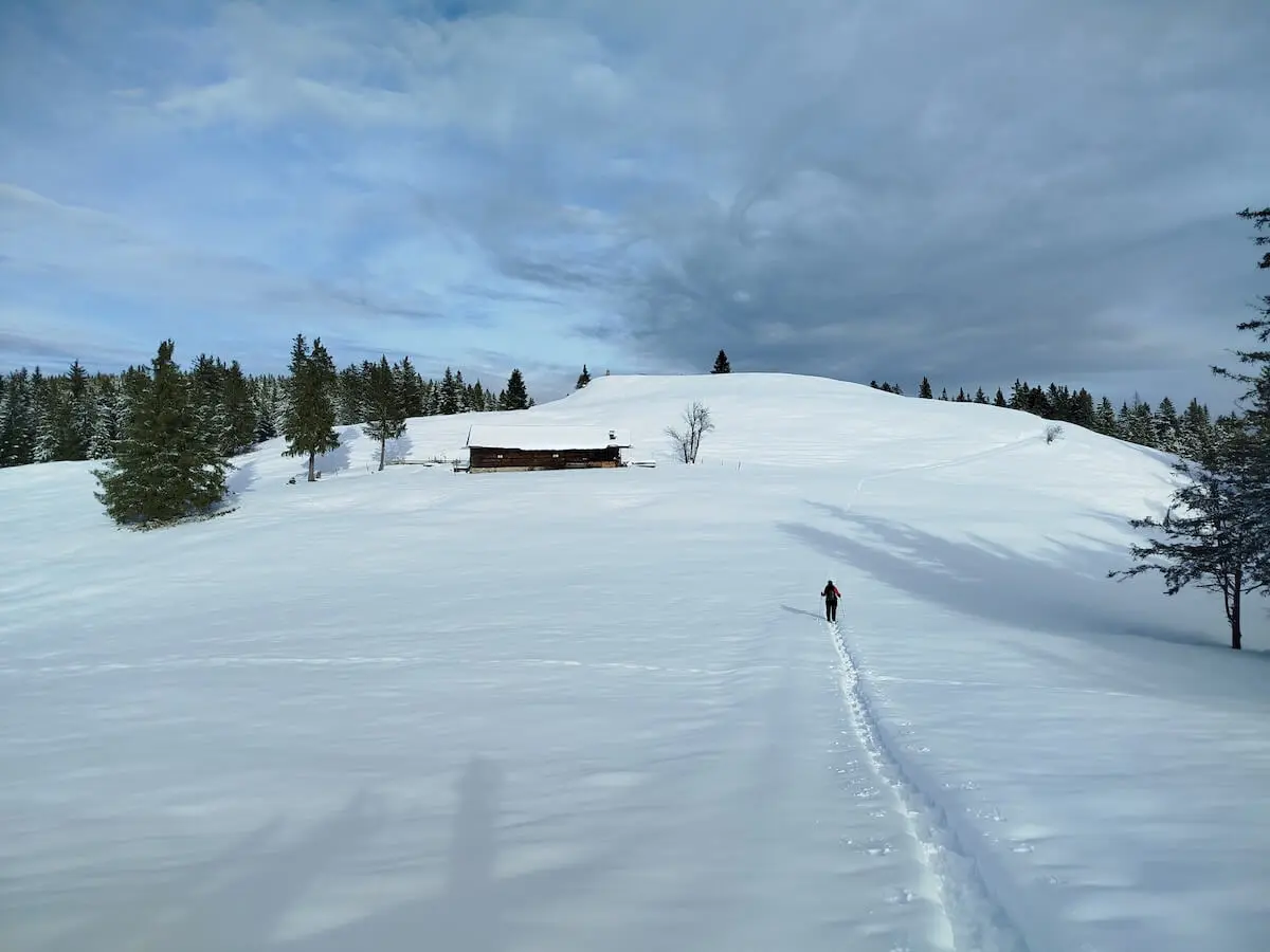 Eine Person wandert durch tiefen Schnee auf eine Holzhütte zu; umgeben von verschneiten Tannen und sanften Hügeln. | © DAV Markt Schwaben | Foto: Christine Strasser