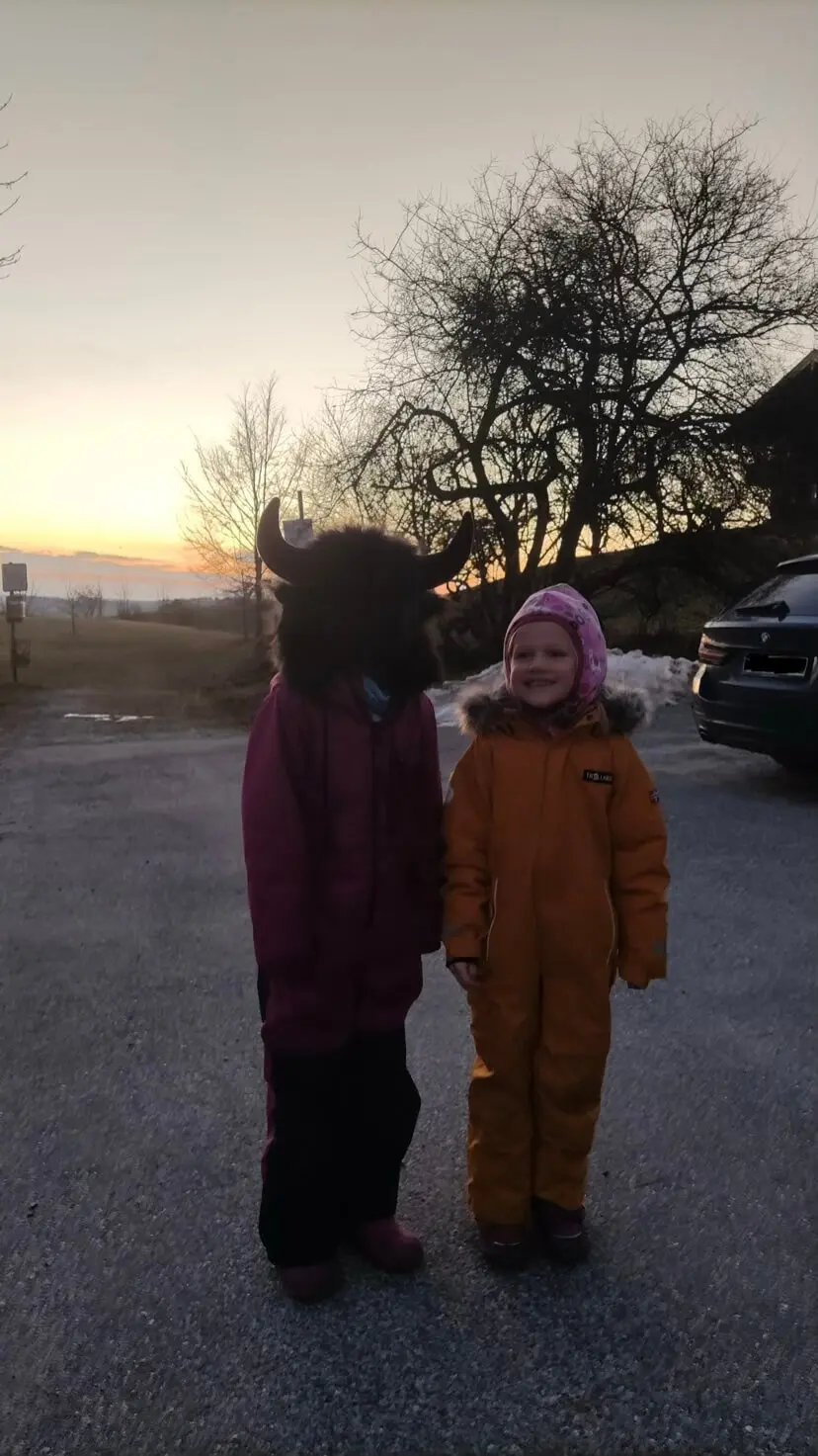 Zwei Kinder im Schneeanzug stehen bei Sonnenuntergang draußen, eines trägt eine Fellmütze mit Hörnern. | © DAV Markt Schwaben Familiengruppe