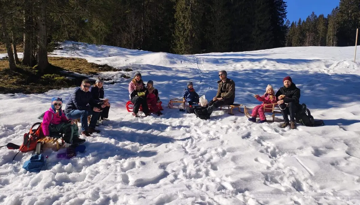 Gruppe von Menschen sitzt im Schnee auf Schlitten und genießt eine Brotzeit, umgeben von hohen Bäumen und winterlicher Stimmung. | © DAV Markt Schwaben