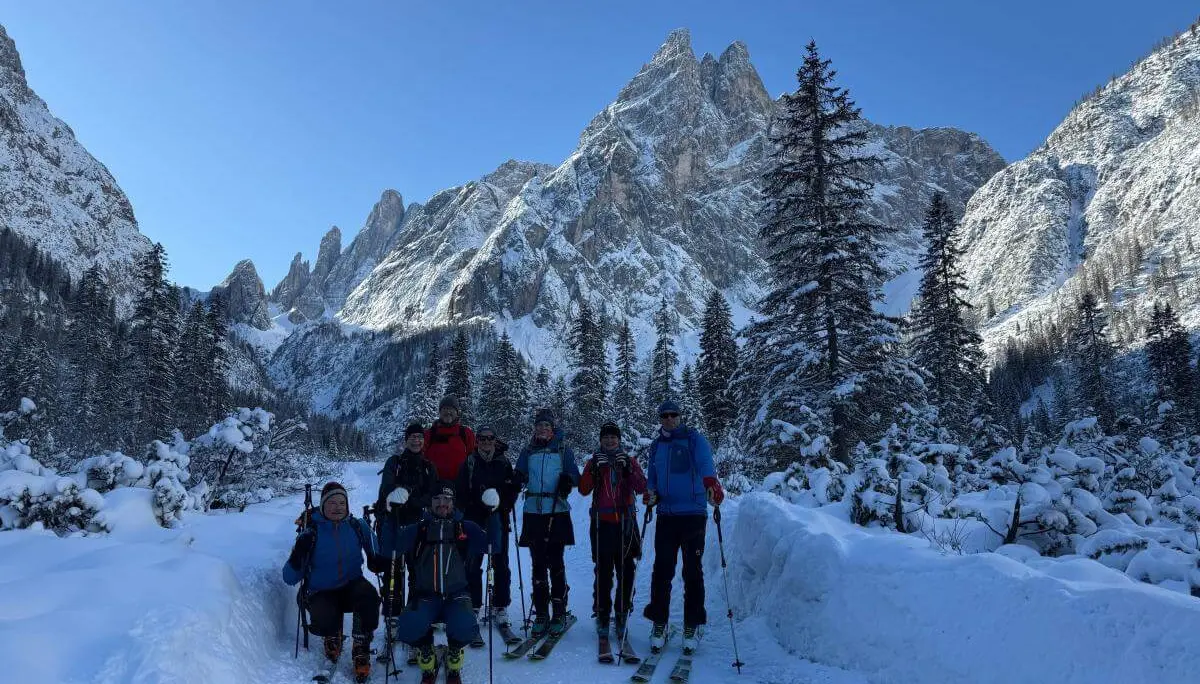 Acht Skitourengeher:innen stehen auf verschneitem Waldpfad im Fischleinbachtal vor sonnenbeschienenen Dolomitengipfeln. | © DAV Markt Schwaben · Foto: Benedikt Scheuerecke