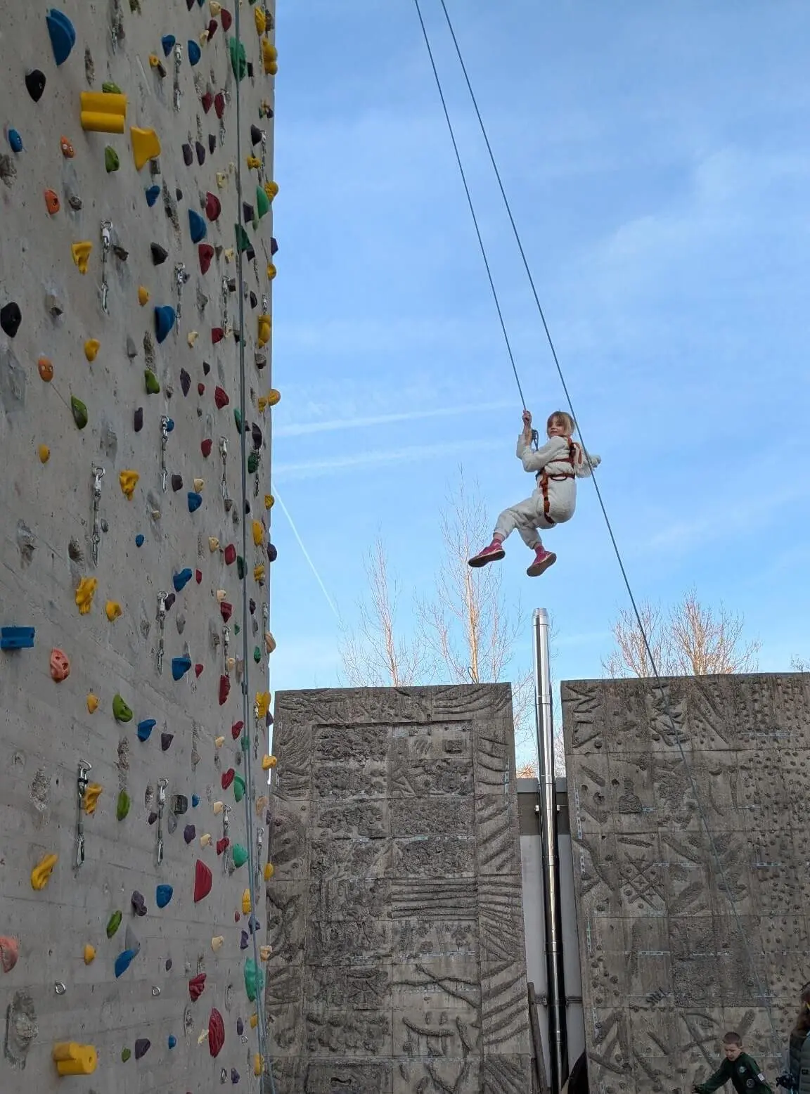 Ein junges Mädchen in einem weißen Overall und rosa Schuhen hängt in der Luft, während es sich von einer hohen Kletterwand im Freien mit bunten Griffen abseilt. | © DAV Markt Schwaben