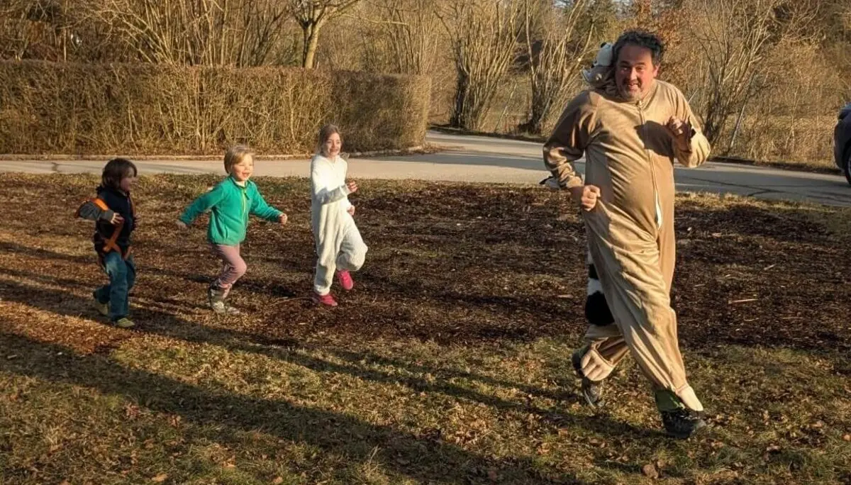 Eine Gruppe von vier Kindern und einem Erwachsenen rennt draußen auf einem mit Laub bedeckten Boden. Der Erwachsene trägt ein Kostüm, das wie ein Tier aussieht. Die Kinder tragen verschiedene farbenfrohe Kleidung. Im Hintergrund sind kahle Bäume und ein Weg zu sehen. Die Szene findet an einem sonnigen Tag statt. | © DAV Markt Schwaben