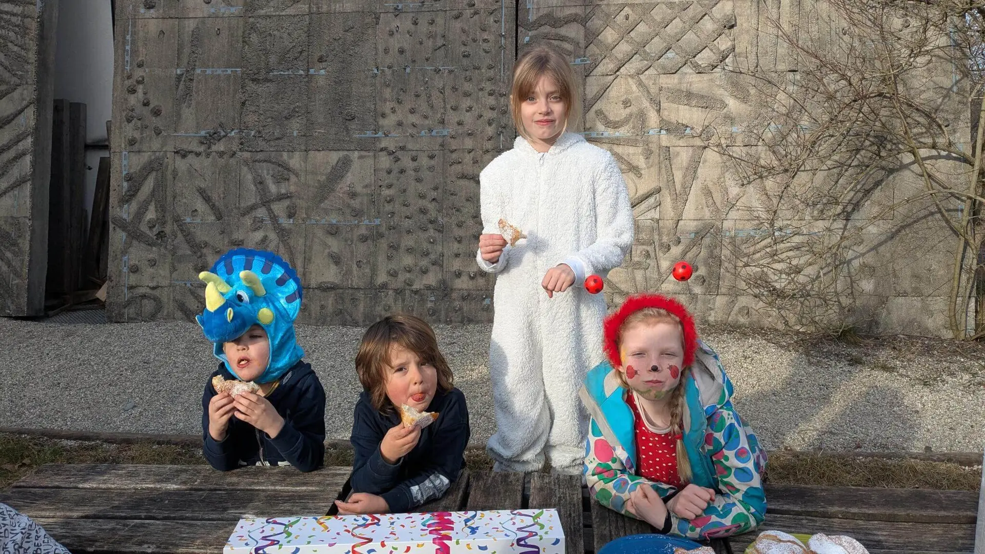 Vier Kinder in bunten und verspielten Outfits sitzen draußen an einem Holztisch und essen Krampfen. | © DAV Markt Schwaben