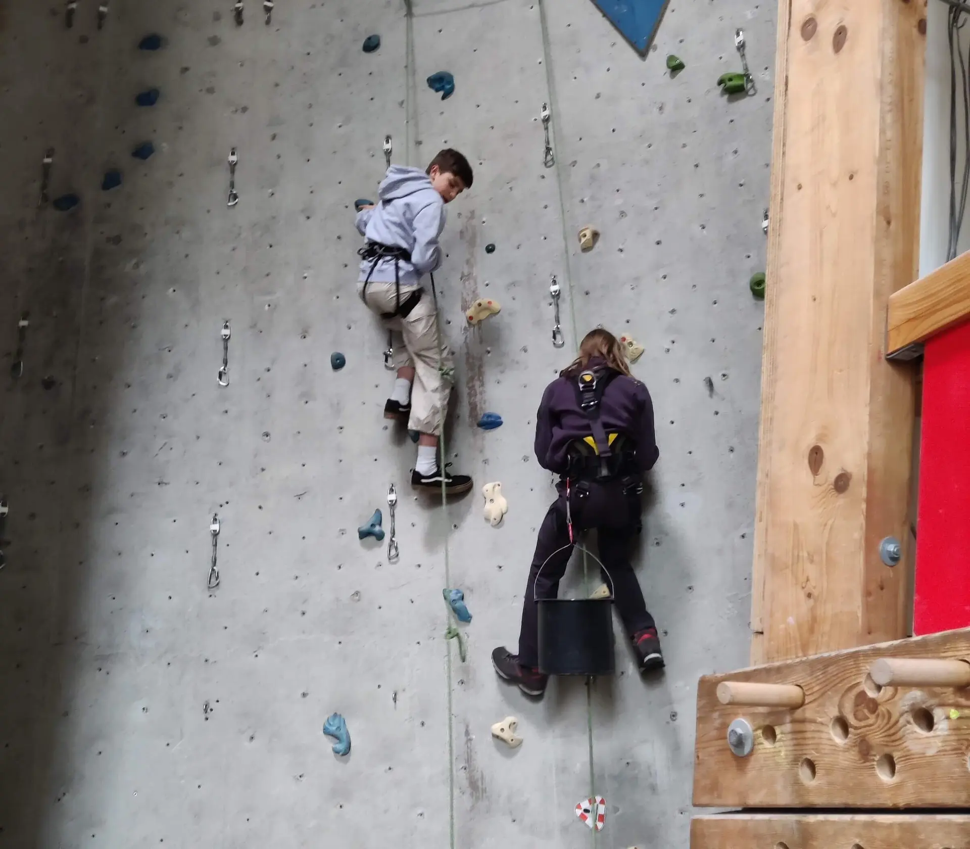 Zwei Personen arbeiten an einer Kletterwand mit bunten Griffen, tragen Sicherheitsgurte und Kletterausrüstung. | © DAV Markt Schwaben