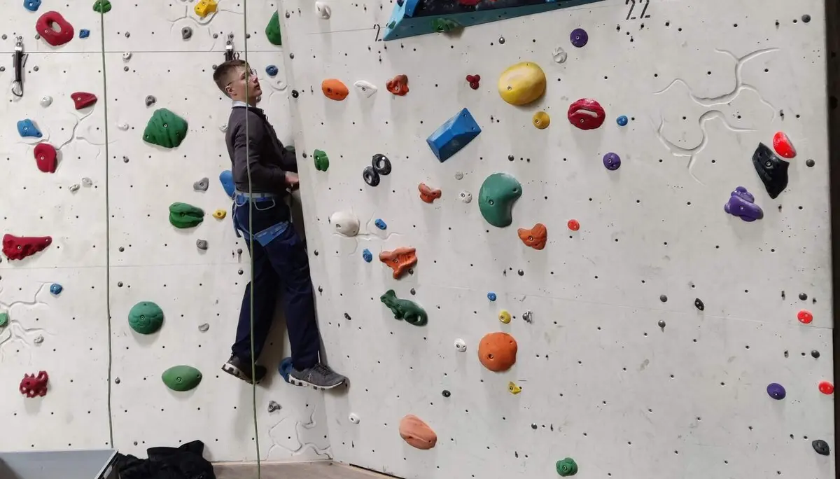 Eine Person klettert gesichert an einer bunten Indoor-Kletterwand, Kletterausrüstung liegt am Boden. | © DAV Markt Schwaben