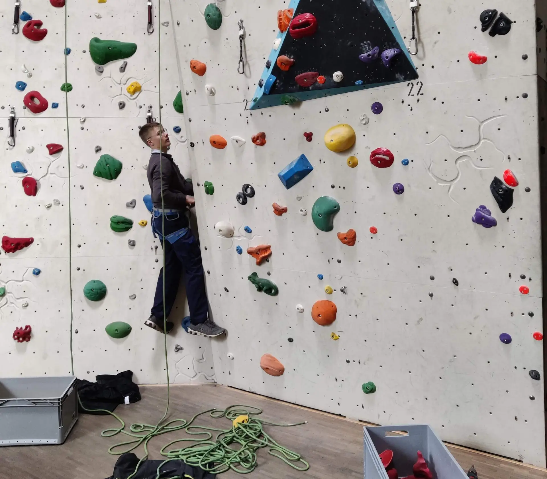 Eine Person klettert gesichert an einer bunten Indoor-Kletterwand, Kletterausrüstung liegt am Boden. | © DAV Markt Schwaben
