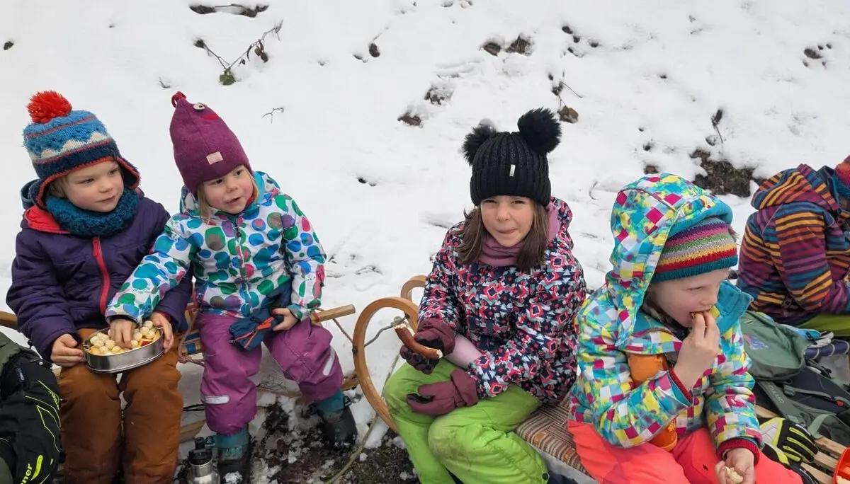 Kinder in bunter Winterkleidung sitzen auf einem Schlitten im Schnee und genießen Snacks aus einem Metallbehälter. | © DAV Markt Schwaben