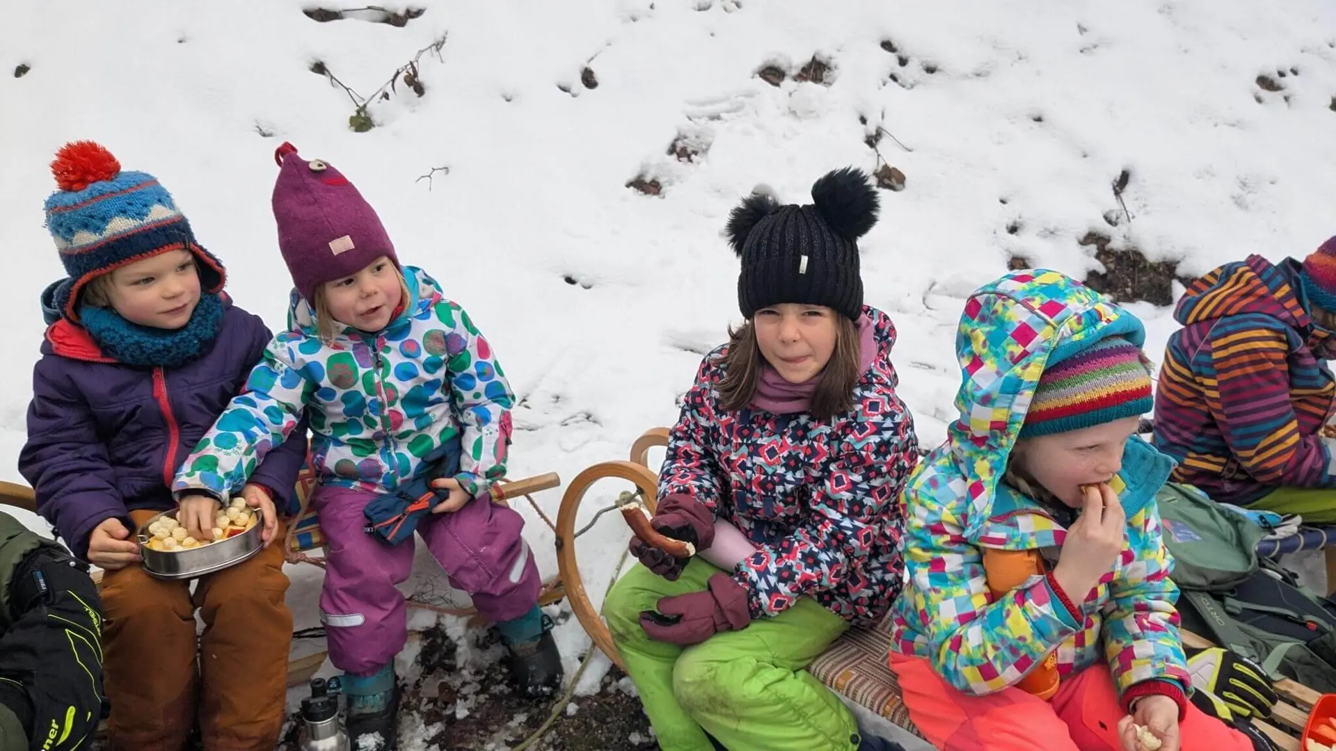 Kinder in bunter Winterkleidung sitzen auf einem Schlitten im Schnee und genießen Snacks aus einem Metallbehälter. | © DAV Markt Schwaben
