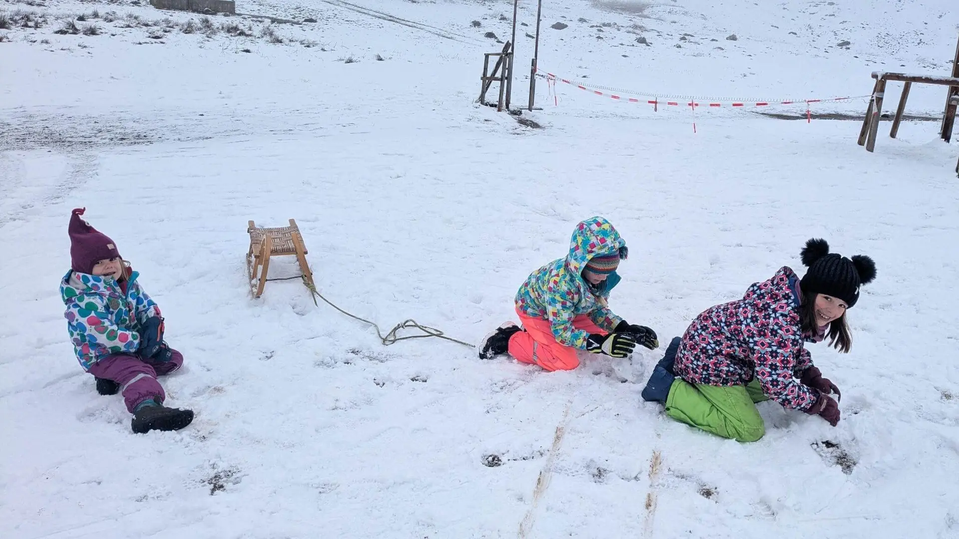 Drei Kinder in bunter Winterkleidung spielen im Schnee mit einem Holzschlitten, umgeben von einer Schneelandschaft.“ | © DAV Markt Schwaben