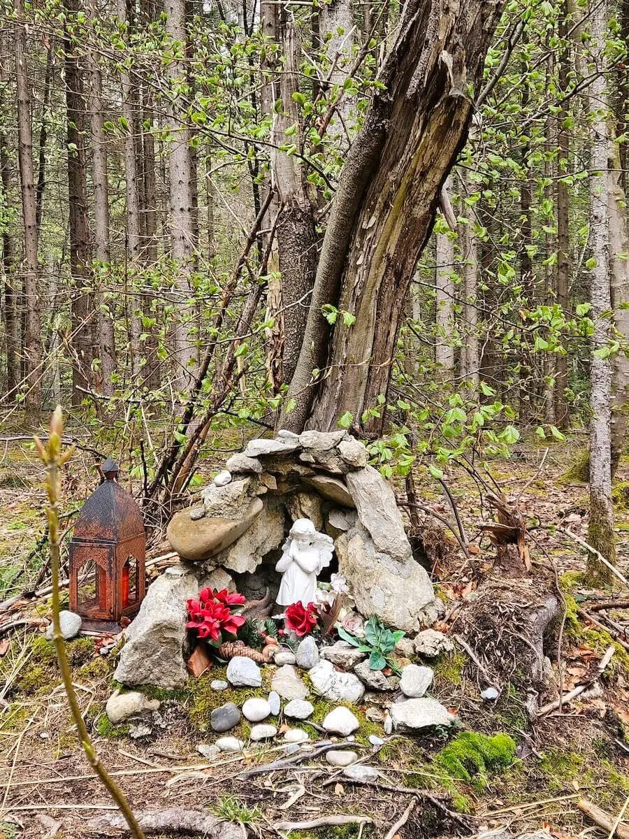 .Gedenkstätte im Wald mit Engelstatue und Laterne. Eine ruhige und besinnliche Szene mit einer Engelstatue, roten Blumen und einer Laterne zwischen Bäumen und Moos. | © DAV Markt Schwaben | Foto Hubert Inhofer