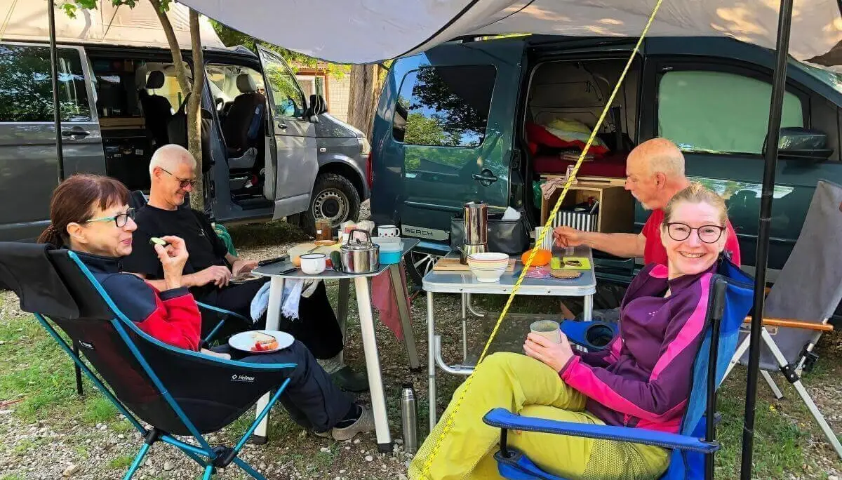 Vier Personen sitzen unter einem Tarp am Campingtisch mit Geschirr und Essen; Campervans im Hintergrund. | © DAV Markt Schwaben | Foto Hildegard Petschik