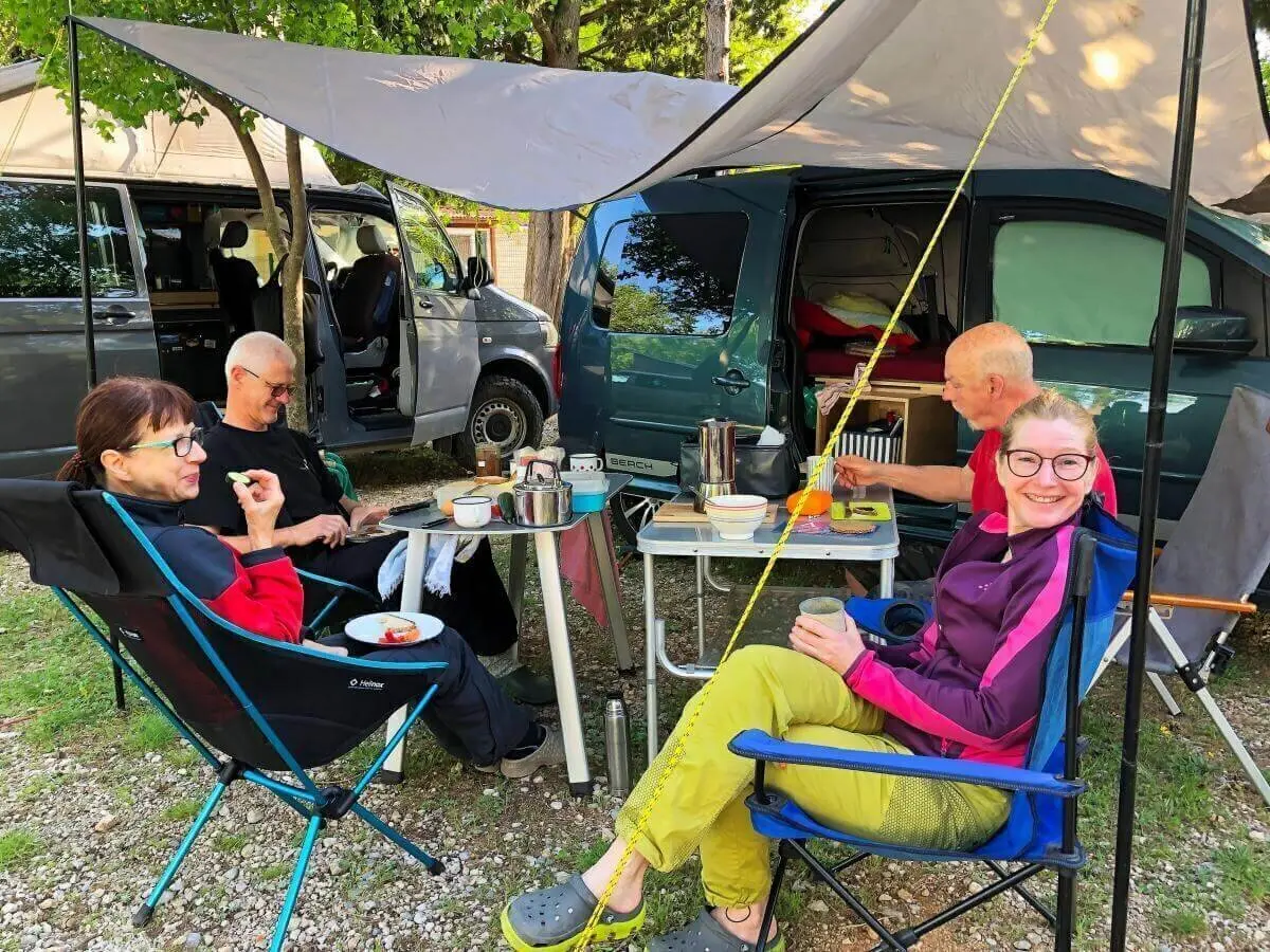Vier Personen sitzen unter einem Tarp am Campingtisch mit Geschirr und Essen; Campervans im Hintergrund. | © DAV Markt Schwaben | Foto Hildegard Petschik