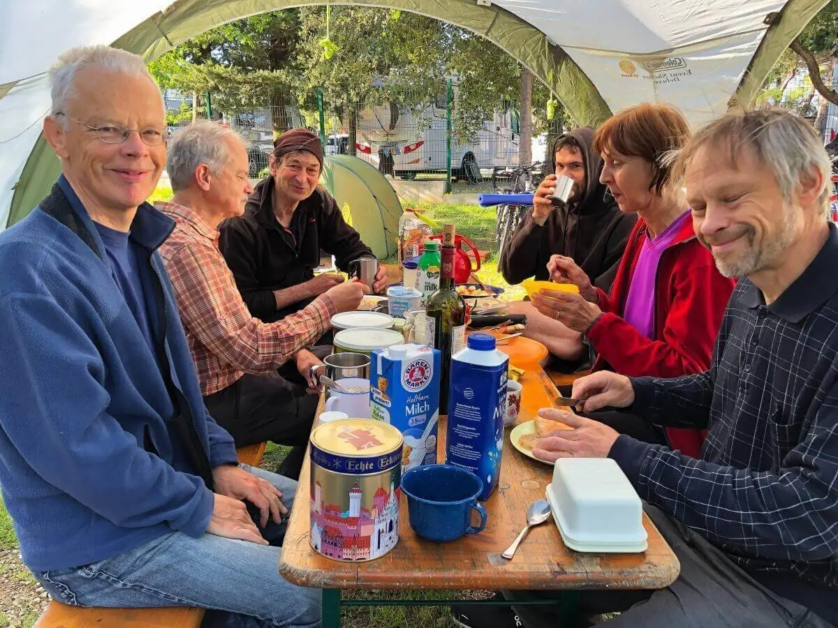Acht Erwachsene sitzen am Picknicktisch unter einem Pavillon, umgeben von Zelten und Bäumen, beim gemeinsamen Frühstück. | © DAV Markt Schwaben | Foto Hildegard Petschik