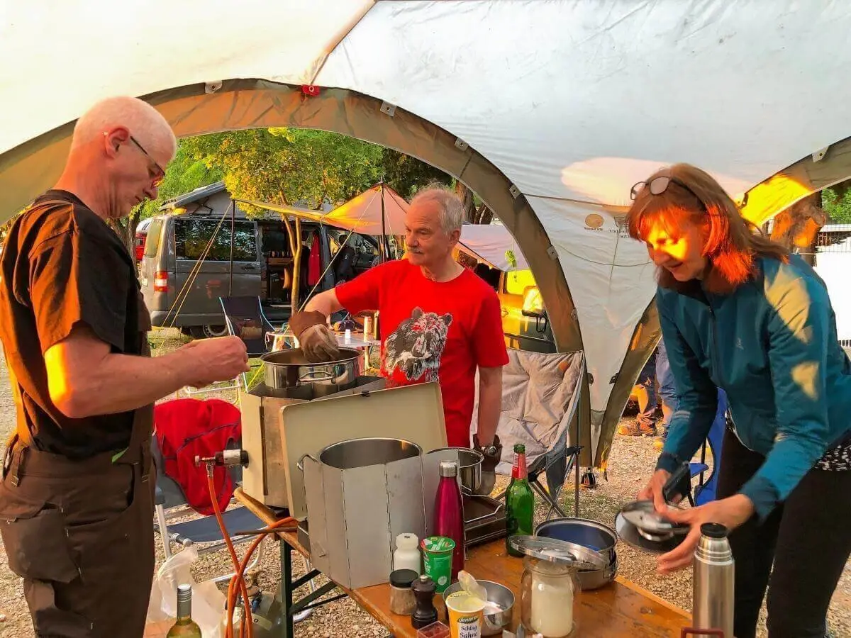 Drei Personen bereiten gemeinsam Essen unter einem Pavillon zu; Campingausrüstung und Zelte im Hintergrund. | © DAV Markt Schwaben | Foto Hildegard Petschik