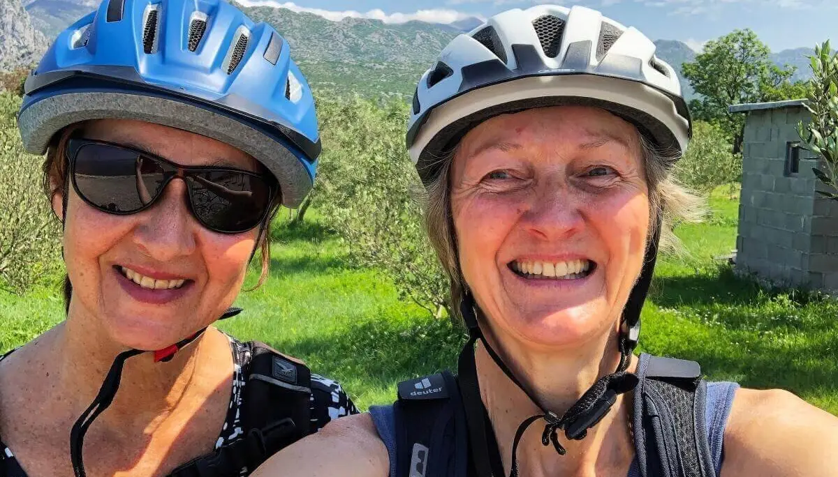 Zwei Personen mit Fahrradhelmen und Rucksäcken stehen auf einer Wiese vor Bergen und bewölktem Himmel. | © DAV Markt Schwaben | Foto Hildegard Petschik